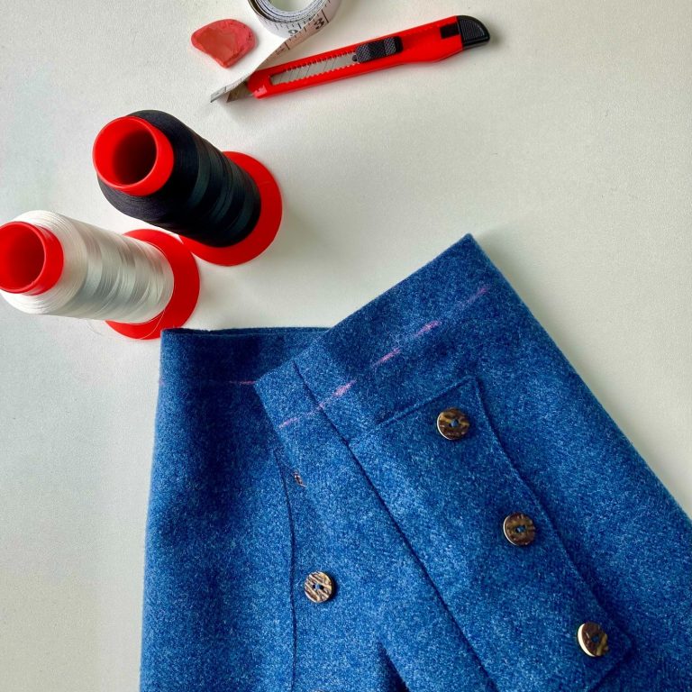 Blue kilt jacket with buttons, thread spools, and a craft knife on a surface.