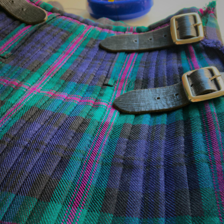 A close-up of a tartan kilt with buckles, featuring green, black, and pink patterns.