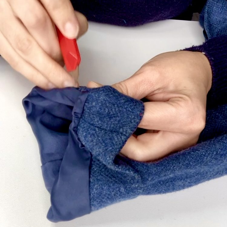 A person's hands adjusting the hem of a blue kilt jacket with a red tool.